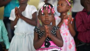 A small girl wears pink and black and holds her hands in prayer. 